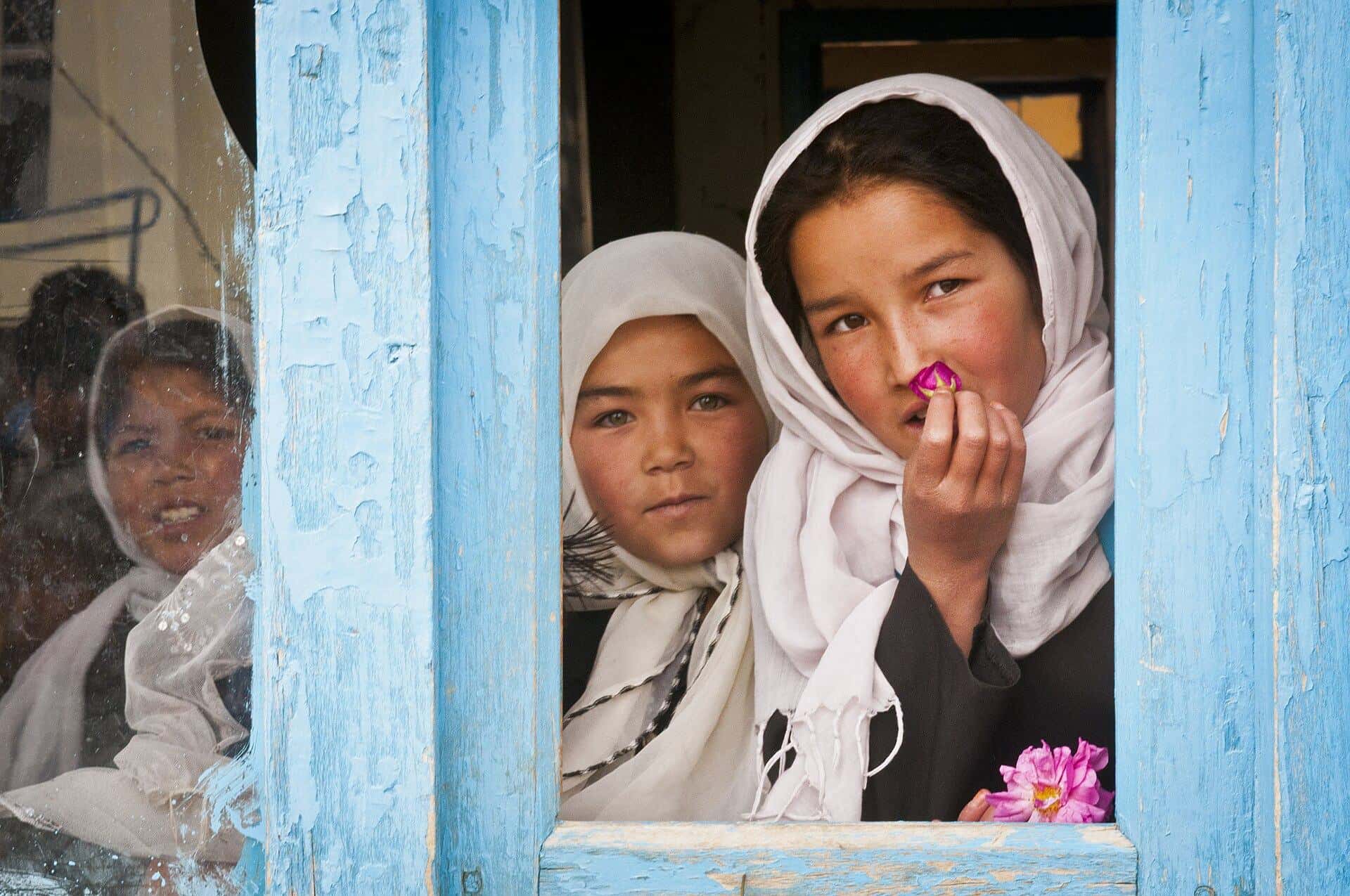 Two schoolgirls in white headscarves look out of a blue-painted classroom window in Bamyan.